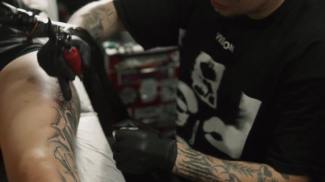Master man tattoo in glasses draws the balck paint on the clients tattoo. Tattoo artist holding tattoo machine in black sterile gloves