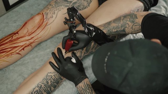 Master man tattoo in glasses draws the balck paint on the clients tattoo. Tattoo artist holding tattoo machine in black sterile gloves