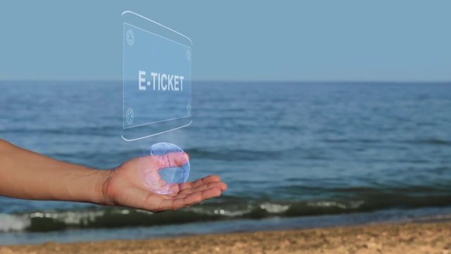 Male hands on the beach hold a conceptual hologram with the text E-ticket. A man with a holographic technology on the background of the sea