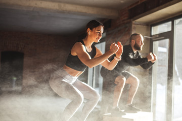 Fit caucasian couple doing squats on boxes in a crossfit style gym, well lit by bright sunlight. - obrazy, fototapety, plakaty