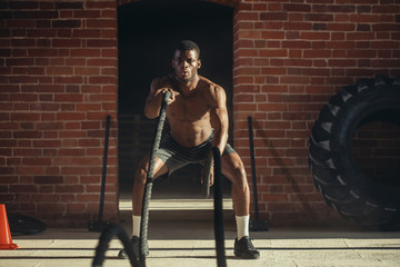Fototapeta na wymiar Hard work of Crossfit instructor of African ethnicity proves that training ropes are a very dynamic workout that accelerates athlete's pulse while strengthening his body.