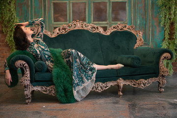 elegant caucasian girl in a luxurious long sequins lace dress with a green fluffy boa in her hands posing in the retro room with vintage luxury furniture