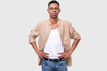 Fototapeta premium Studio portrait of young African American man wearing casual white t-shirt and beige shirt, looking to the camera through round eyeglasses and posing over white background. People emotion concept
