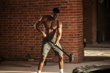Muscular african american crossfit man with naked torso hitting wheel tire with sledgehammer during functional workout in brick wall interior gym