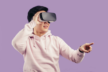 Korean young man surprised by something in virtual reality, wearing virtual reality glasses showing gesture isolated on studio shot on violet wall background