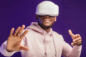 African American guy in white hoodie is playing a virtual reality game using head-mounted display, feeling excitement and joy, isolated over purple background with copyspace