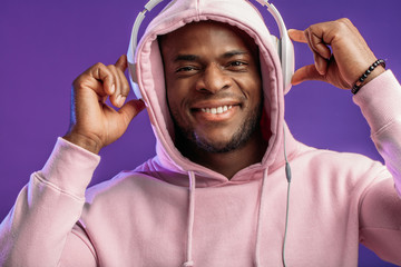 Cute African-American man in headphones dressed in street style wear, listening to music on violet background
