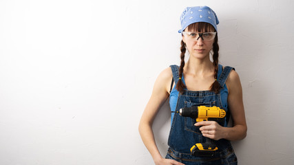 Girl and male tools: a young girl with blue clothes and goggles is standing against a white wall and holding a yellow screwdriver.