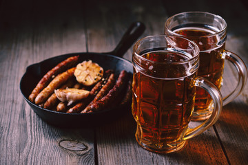 beer and appetizing snacks. table with two mugs of craft lager and frying pan with homemade grilled sausages. oktoberfest food, pub concept