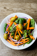Freshly made vegetable salad with carrot, olives, spinach and onion in a bowl on wooden background.