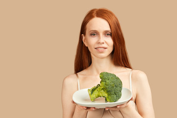 Young skinny woman with ginger hair and starved face in underwear looking at camera holding plate with raw broccoli, happy to eat something