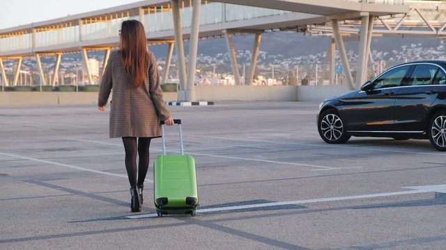 Back view of young business woman with suitcase going to luxury black car.