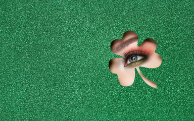 Conceptual photo of St. Patrick's Day. The Eye of the Young Beautiful Woman with Bright Golden Shadows and Expressive Eyebrows, Looks in the Shamrock Leaves Pattern out of Green Paper