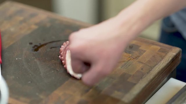 Close up of male hands cutting the initial part of a fresh purple octopus tentacle, with a kitchen knife, on a wooden cutting board, while someone is explaining what to do, in a professional kitchen