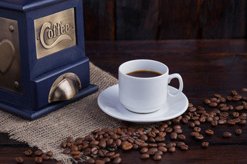 White cup of coffee next to a coffee grinder and ripe coffee grains on a dark wooden background with linen cloth