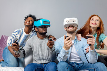 Two multiracial young couples having fun while visiting entertainment center in weekend. Emotional guys in VR headsets experience joy and excitement while driving virtual race car