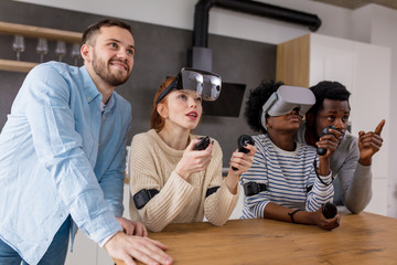 Multicultural group of friends playing video games wearing virtual reality glasses with controllers, women trying to play and boyfriends making hints