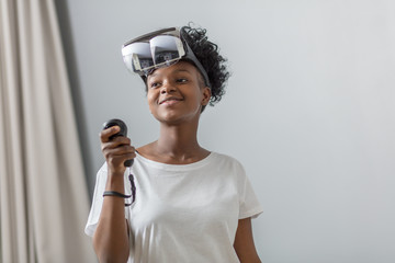 Mixed-race african woman with short curly hair and pleasant smile wears virtual reality goggles with insert smartphone, holds game controllers, standing at home