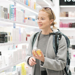 Blond young female traveler wearing coat and travel backpack choosing perfume in airport duty free...