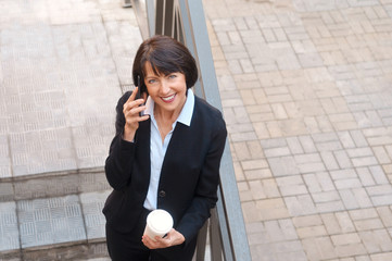 Mature businesswoman having a break in front of an office building, talking on the phone and drinking coffee