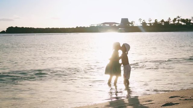 Two little happy kids are standing on the beach near the sea, holding hands and kissing each other. Brother and sister. Slow motion