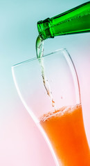 A bottle of green glass pouring beer into a beer glass. At an angle. Turquoise pink background. Close-up.