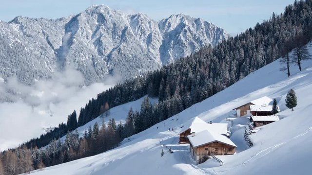 Beautiful 4K timelapse of the sun shining on an Austrian farmhouse in winter. Skiers are going by and the fog and clouds move up and down the small valley in front of jagged, snowy mountain peaks.