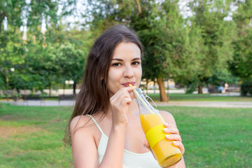 Attractive girl is drinking fresh juice through the straw outdoors. Pretty woman is holding a bottle of cold lemonade
