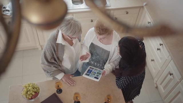 Senior woman showing to her friends photos on tablet. Group of three middle aged mature women communicating chatting spending time in the kitchen at home with wine and fruits. Slow motion. Top view