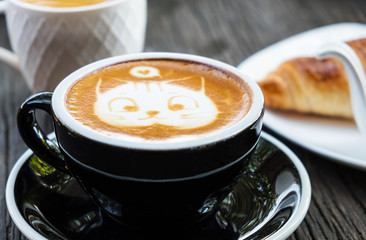 Cat face design of latte art coffee in black cup on old wooden table , there is croissant on white plate in the background