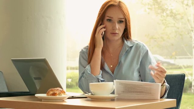 Angry, young businesswoman talking on cellphone with documents in cafe, 4K 