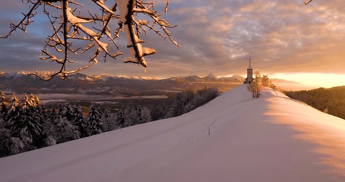 Right side pan of a church on a hill on a snow filled landscape, then revealing a lovely sunrise orange sun with a valley beneath.