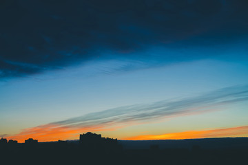 Cityscape with wonderful varicolored vivid dawn. Amazing dramatic blue cloud sky above dark silhouettes of city building roofs. Atmospheric background of orange sunrise in overcast weather. Copy space