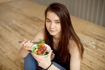 Sports beautiful girl sitting on the floor and eating a salad from fresh vegetables and greens. Healthy lifestyle.