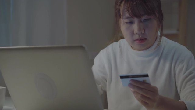 Beautiful Asian woman using laptop buying online shopping by credit card while wear casual sitting on desk in living room in the night at home. Lifestyle women working at home office concept.
