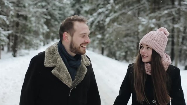 Young attractive couple walking and having fun in a winter forest under heavy snow. Man and woman in pink hat hugging and holding hands toghether. Happy family in winter forest. Slow motion.