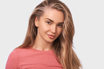 Fototapeta premium Studio close up portrait of blonde good looking young woman, with beautiful healthy hair, smiles positively wearing casual outfit and looking to the camera, isolated on white wall. People emotions