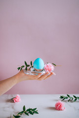 The girl is holding a blue eater egg on a stand, pink and marble background, minimalism, flowers