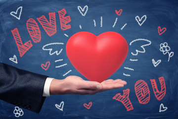 Male hand holding red heart with LOVE YOU chalk sign and heart drawings on blue background