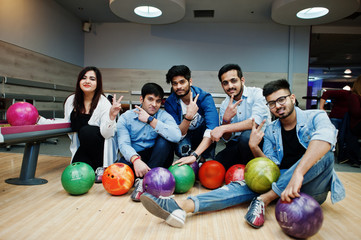 Group of five south asian peoples having rest and fun at bowling club. Holding bowling balls, sitting on alley and show two fingers sign.