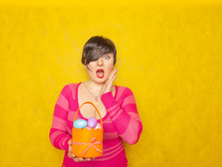 pretty adult chubby woman with short hair holding colorful holiday eggs and a easter basket on yellow solid studio background