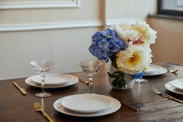 white plates, knifes and forks, crystal glasses and flowers in vase on wooden table at home