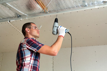Young man in goggles fixing drywall suspended ceiling to metal frame using electrical screwdriver...