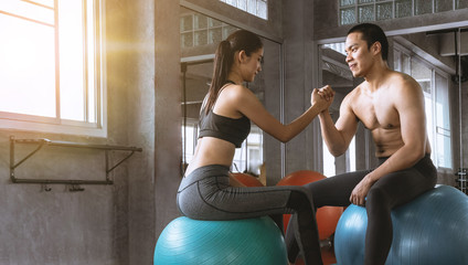 Muscular Asian couple giving hand shake while sitting on yoga ball at the gym. Happy work out and exercise concept.