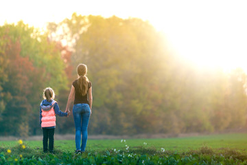 Back view of young slim attractive mother and child girl standing in green meadow holding hands outdoors on forest trees blurred background.