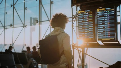 Funny casual looking and authentic young traveller with curly crazy hair studies flight information display in departure terminal of international or domestic airport, travel blogger inspiration