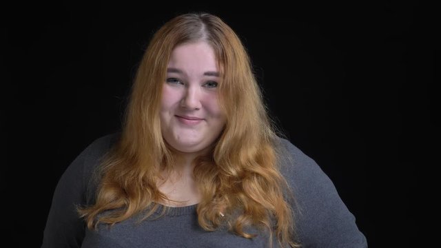 Closeup shoot of young overweight caucasian female being shy and looking at camera smiling with background isolated on black