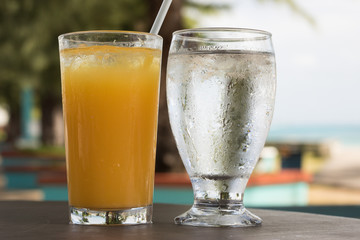 Close-up: Glass of Orange Juice Ice Cocktail and Another Glass of Cold Water are on a Wooden Table Against Background of Summer Nature Outside. Cold Drinks are on Hot Day. 