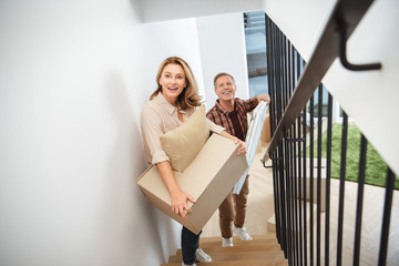 cheerful couple carrying things upstairs and looking at camera
