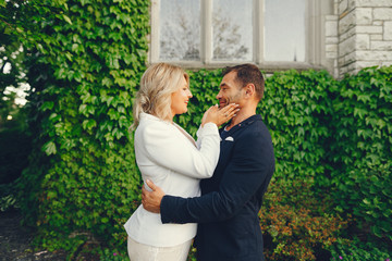 elegant and stylish bride with light curly hair dressed in a white dress, a white jacket and white dress standing in a summer garden on the background of old big houses along with her elegant man